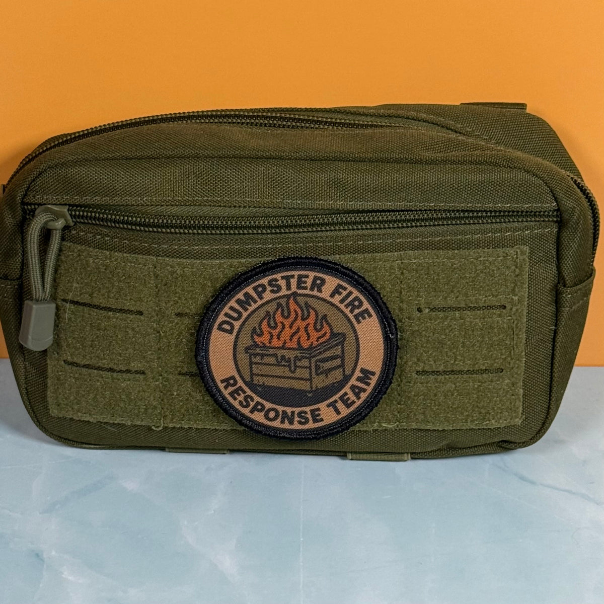 An olive green tactical pouch with a circular 'Dumpster Fire Response Team' patch on the front, set against a split orange and light blue background.