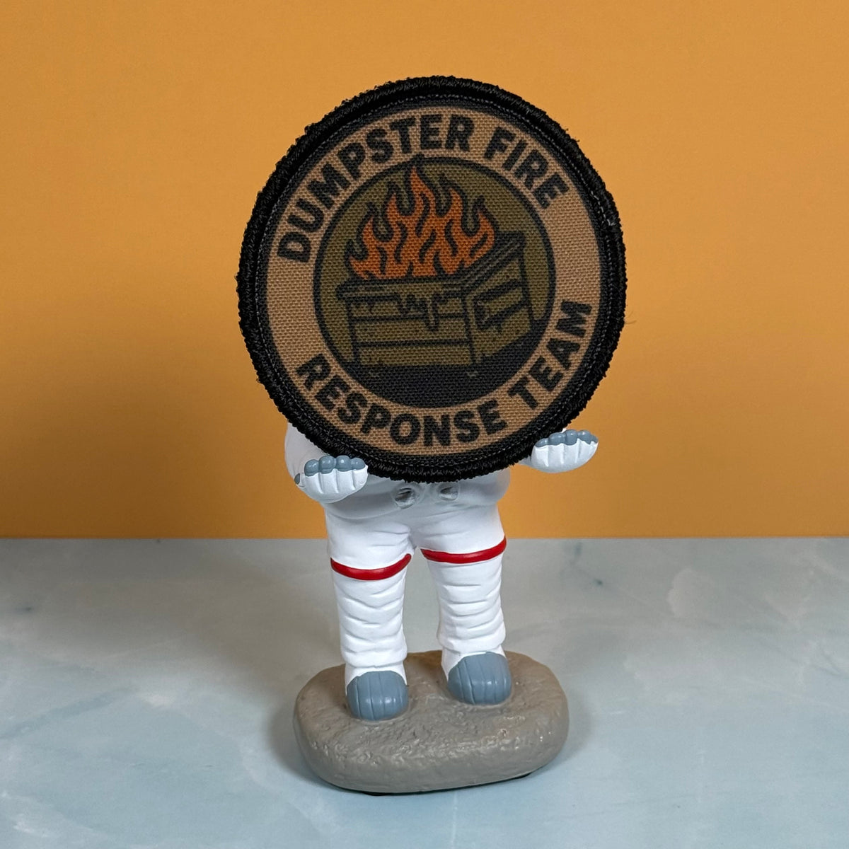 A circular embroidered patch reading 'DUMPSTER FIRE RESPONSE TEAM' with a fire graphic, held by a small white astronaut figurine on a gray base against an orange wall and pale surface.