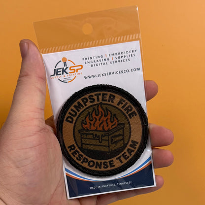 A hand holds a packaged circular embroidered patch that reads "DUMPSTER FIRE RESPONSE TEAM" with a flame and dumpster graphic. The card backing shows the Jeksp brand and service details, against a solid orange background.