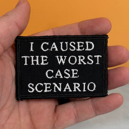 A black rectangular embroidered patch with white text reading 'I CAUSED THE WORST CASE SCENARIO', held in a person's hand against a solid orange background.