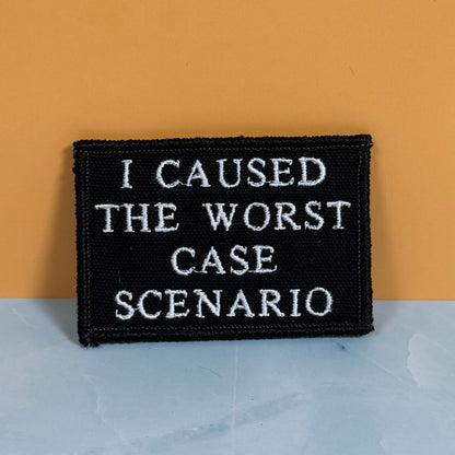 A rectangular black embroidered patch with white text reading 'I CAUSED THE WORST CASE SCENARIO', photographed against a two-tone background with a mustard yellow upper section and a pale blue lower section.