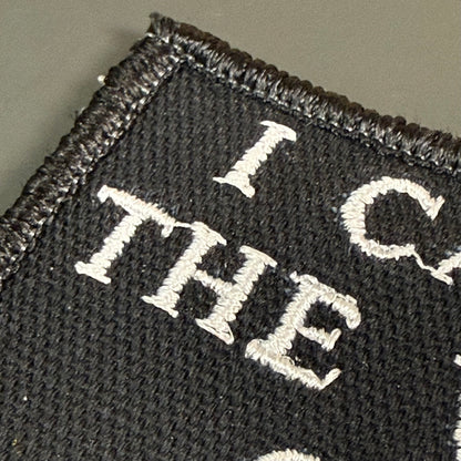Close-up of a black embroidered patch with white lettering reading 'I CAN'T' on a dark textured fabric background.
