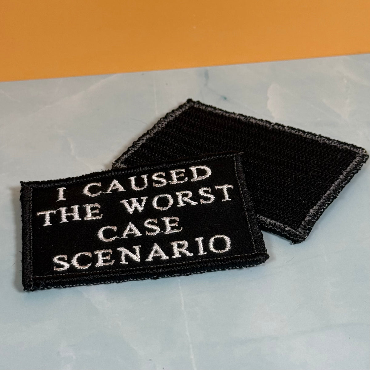 Two rectangular black embroidered patches with white text reading 'I CAUSED THE WORST CASE SCENARIO,' shown on a light marble-like surface with an orange background.