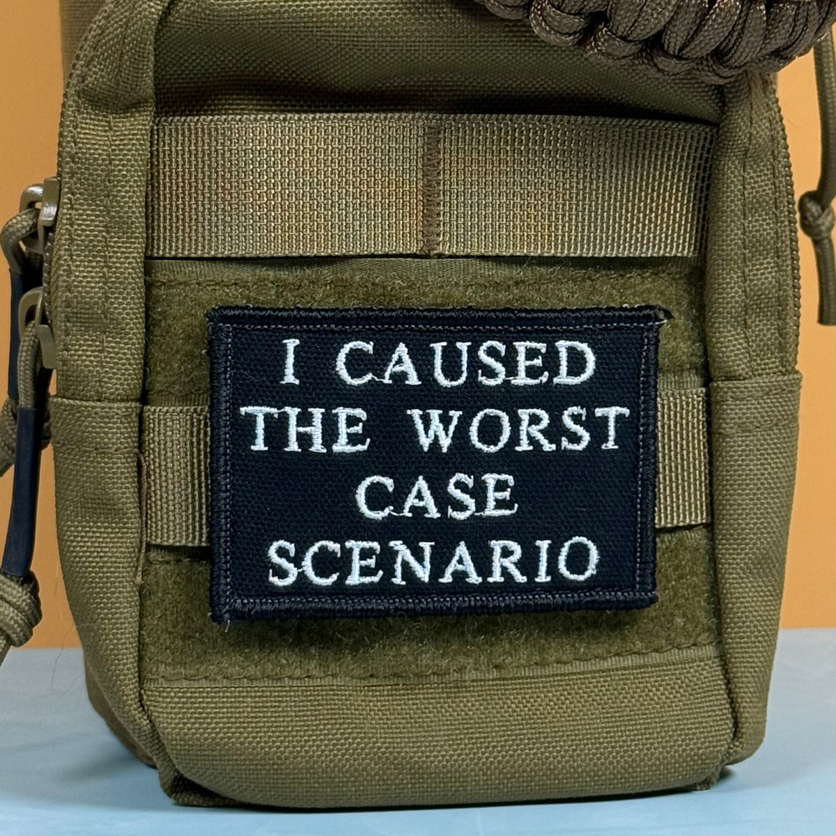 An olive green tactical pouch with a black rectangular embroidered patch that reads 'I CAUSED THE WORST CASE SCENARIO' in white text, shown against a plain background.