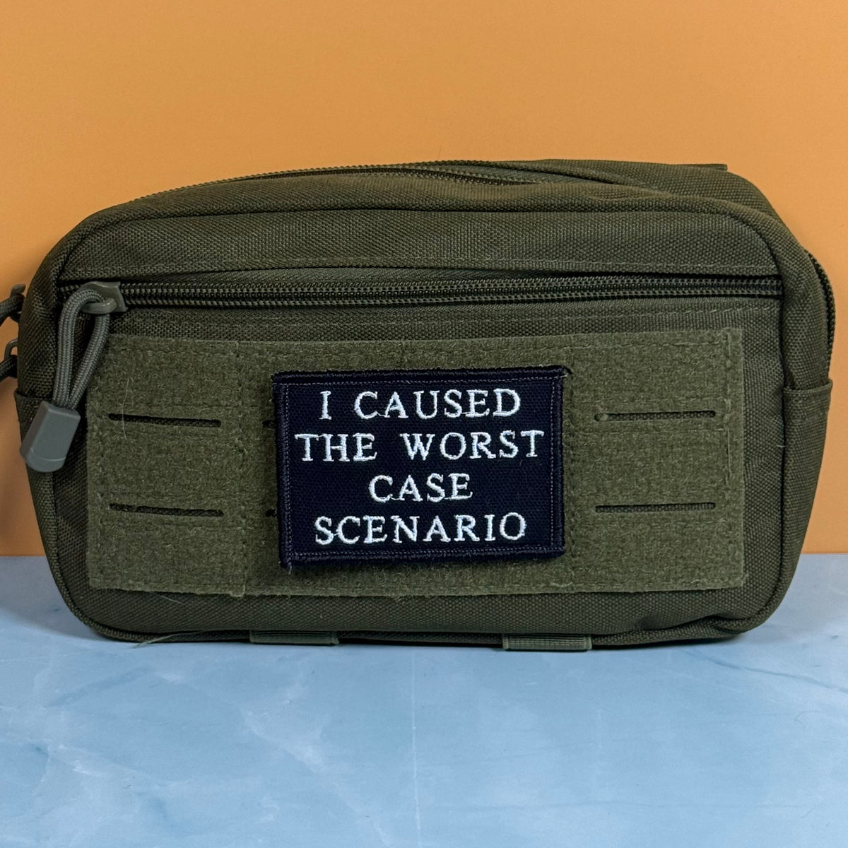 An olive green tactical pouch with a black rectangular patch that reads 'I CAUSED THE WORST CASE SCENARIO' in white text, set against a split orange and light blue background.
