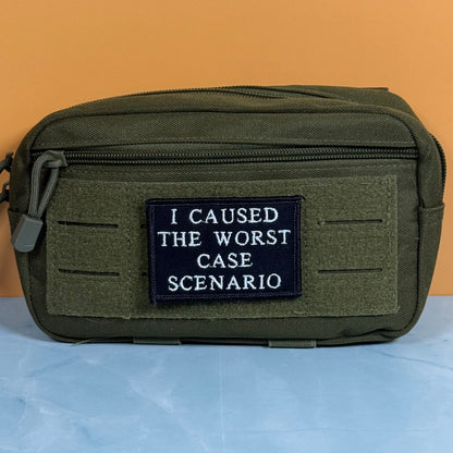 An olive green tactical pouch with a black rectangular patch that reads 'I CAUSED THE WORST CASE SCENARIO' in white text, set against a split orange and light blue background.
