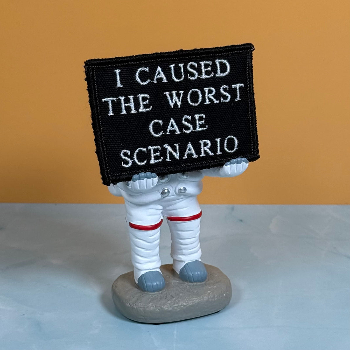 A small astronaut figurine holding a black rectangular patch with white embroidered text that reads 'I CAUSED THE WORST CASE SCENARIO.' The astronaut is white with red bands and gray boots, standing on a gray base against a warm tan background and pale blue surface.
