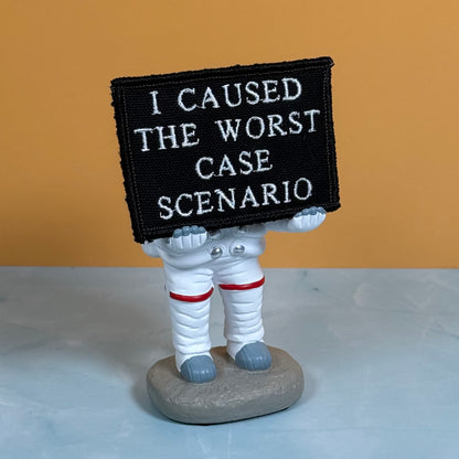 A small astronaut figurine holding a black rectangular patch with white embroidered text that reads 'I CAUSED THE WORST CASE SCENARIO.' The astronaut is white with red bands and gray boots, standing on a gray base against a warm tan background and pale blue surface.