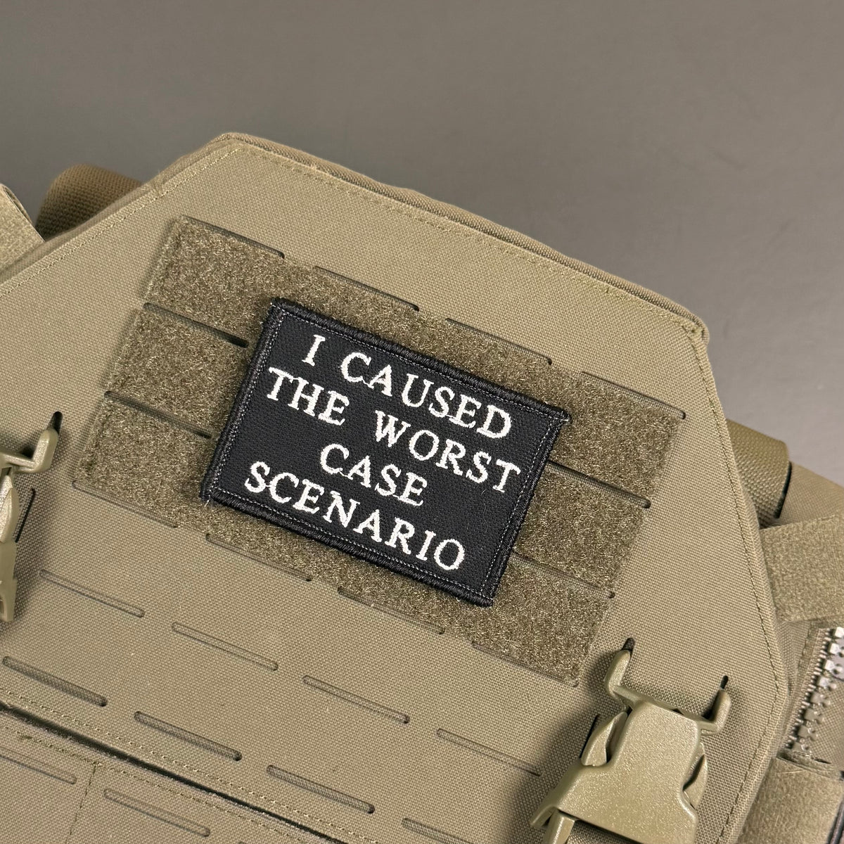 A close-up of a tactical patch with the text 'I CAUSED THE WORST CASE SCENARIO' in white on a black background, attached to a tan MOLLE webbing panel on a tactical vest or backpack.