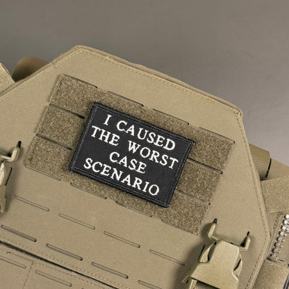 A close-up of a tactical patch with the text 'I CAUSED THE WORST CASE SCENARIO' in white on a black background, attached to a tan MOLLE webbing panel on a tactical vest or backpack.