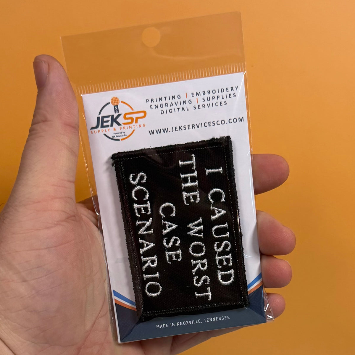 A hand holds a clear plastic blister pack containing a black embroidered patch with white text reading 'I CAUSED THE WORST CASE SCENARIO.' The backing card shows the Jeksp Digital Printing logo and website, against a solid orange background.