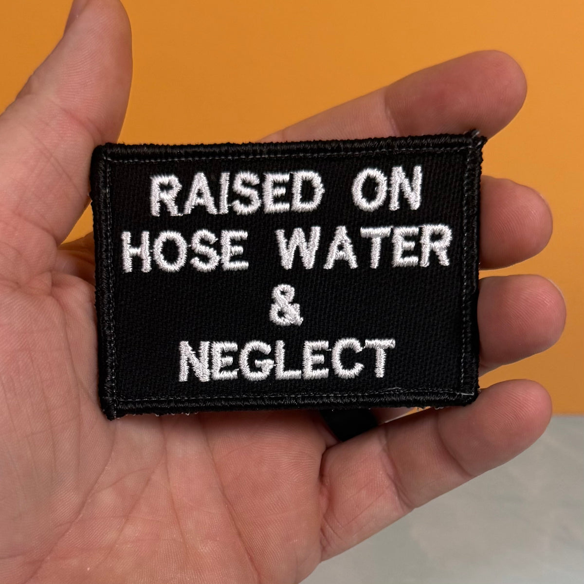 Raised on Hose Water & Neglect embroidered morale patch: hand holding a rectangular black embroidered patch with white text that reads 'RAISED ON HOSE WATER & NEGLECT' against an orange background.