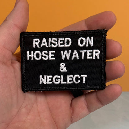 Raised on Hose Water & Neglect embroidered morale patch: hand holding a rectangular black embroidered patch with white text that reads 'RAISED ON HOSE WATER & NEGLECT' against an orange background.