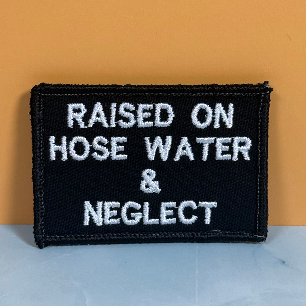 Raised on Hose Water & Neglect embroidered morale patch: black embroidered patch with white text reading 'RAISED ON HOSE WATER & NEGLECT' against a warm orange background and light gray surface.