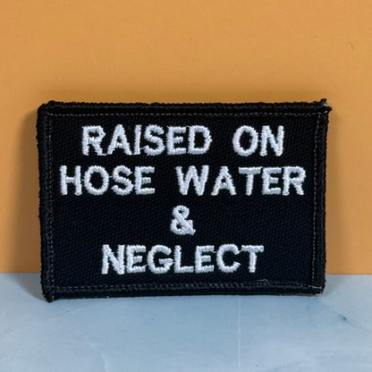 Raised on Hose Water & Neglect embroidered morale patch: black embroidered patch with white text reading 'RAISED ON HOSE WATER & NEGLECT' against a warm orange background and light gray surface.
