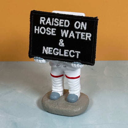 Raised on Hose Water & Neglect embroidered morale patch: astronaut figurine holding a black rectangular sign with white embroidered text that reads 'RAISED ON HOSE WATER & NEGLECT'. The astronaut is dressed in a white spacesuit.