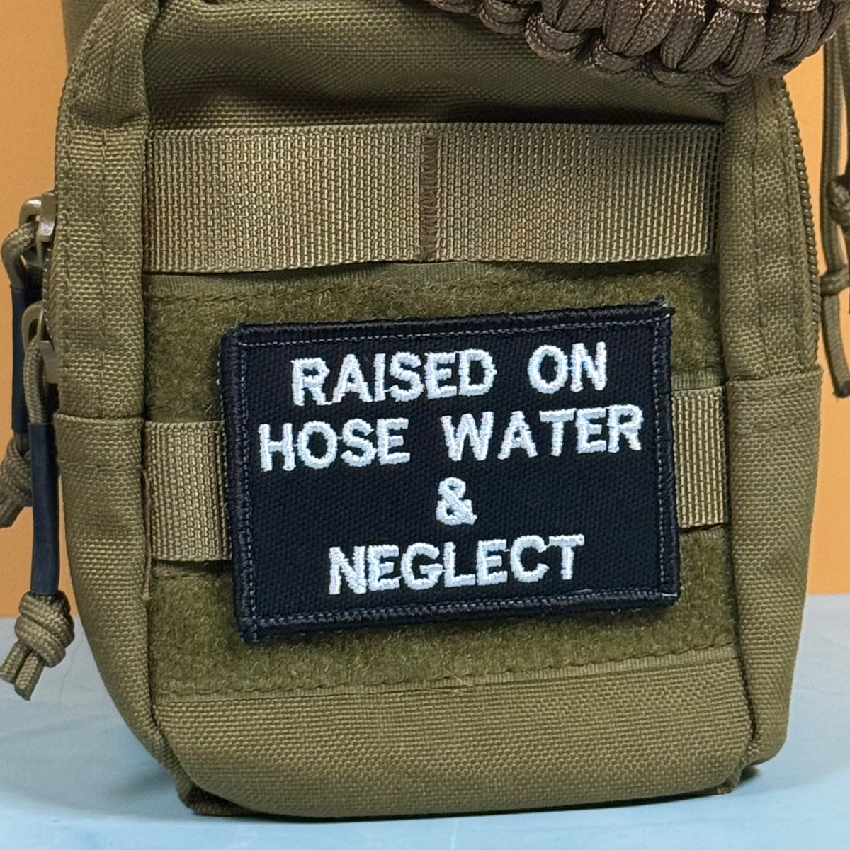 Raised on Hose Water & Neglect embroidered morale patch: olive green tactical pouch with a black rectangular patch that reads 'RAISED ON HOSE WATER & NEGLECT' in white embroidered text.