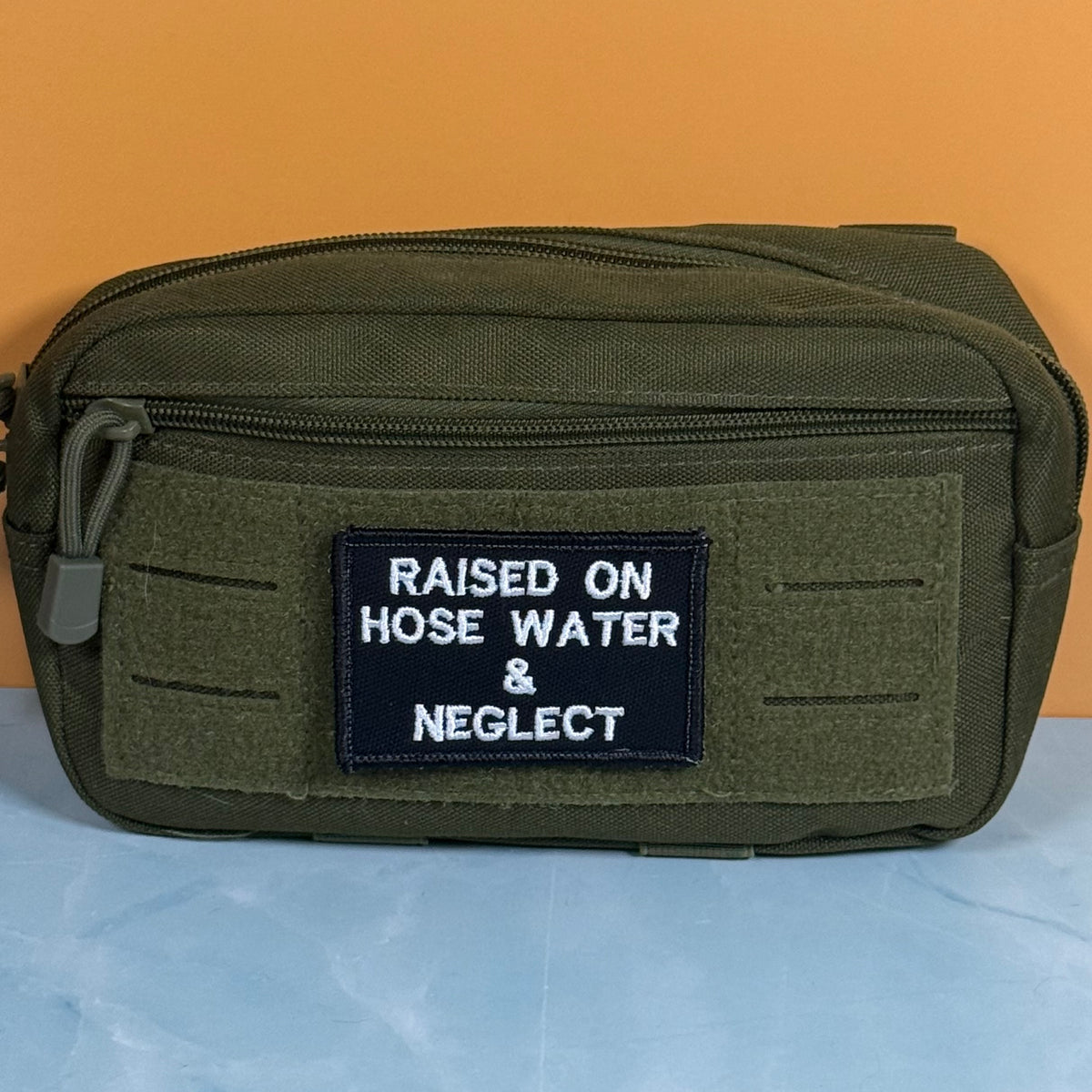 Raised on Hose Water & Neglect embroidered morale patch: olive green tactical pouch with a black embroidered patch that reads 'RAISED ON HOSE WATER & NEGLECT' in white text, set against a split orange and light blue background.