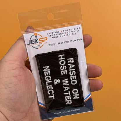 Raised on Hose Water & Neglect embroidered morale patch: hand holding a packaged embroidered patch that reads 'RAISED ON HOSE WATER & NEGLECT' in white lettering on a black background. The patch is inside a clear plastic sleeve.
