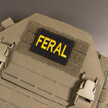 A close-up of a tactical plate carrier vest in coyote tan, featuring a black rectangular patch with the word 'FERAL' embroidered in bright yellow.