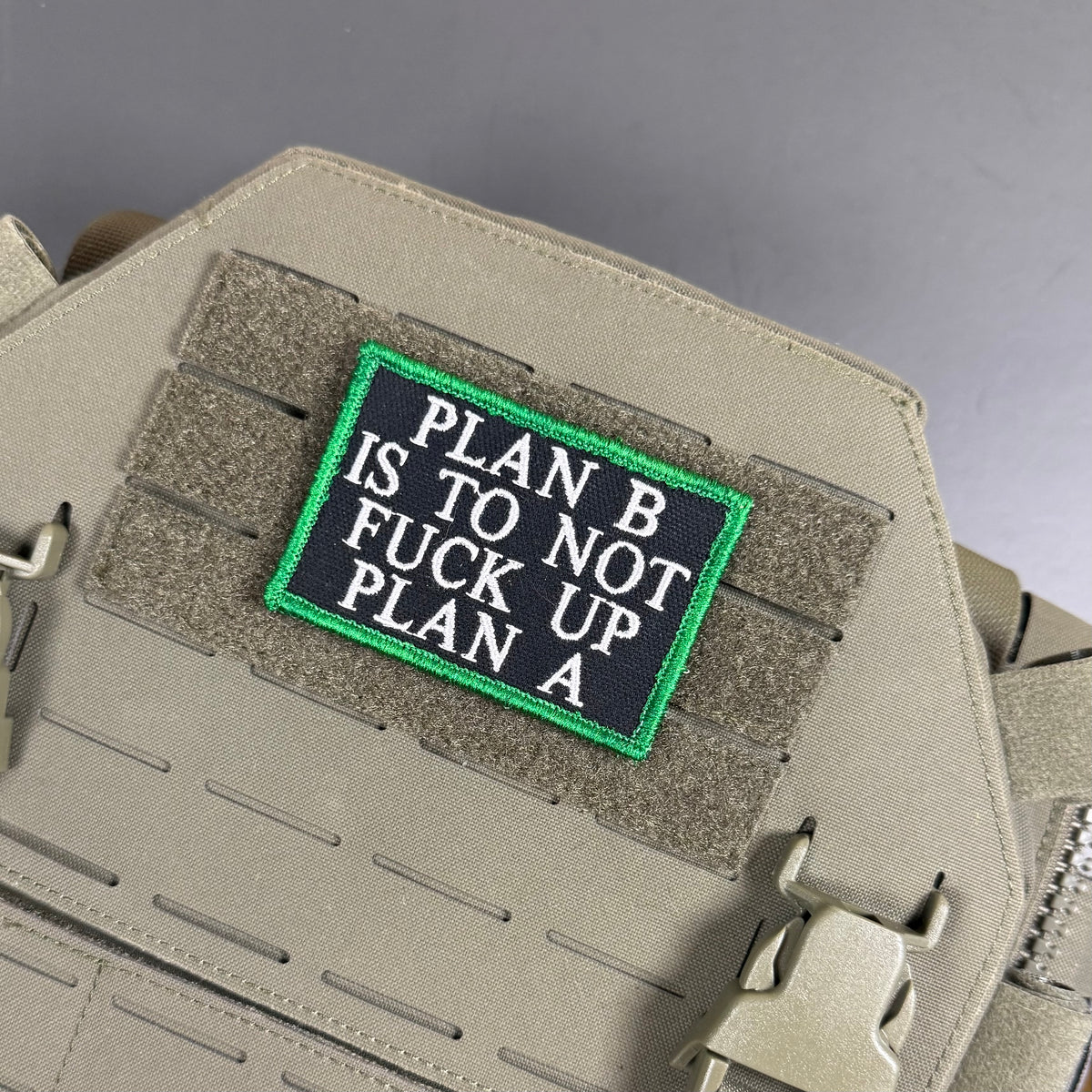 A rectangular embroidered tactical patch with the text 'PLAN B IS TO FUCK UP PLAN A' in white letters on a black background with a bright green border, shown attached to tan tactical gear.