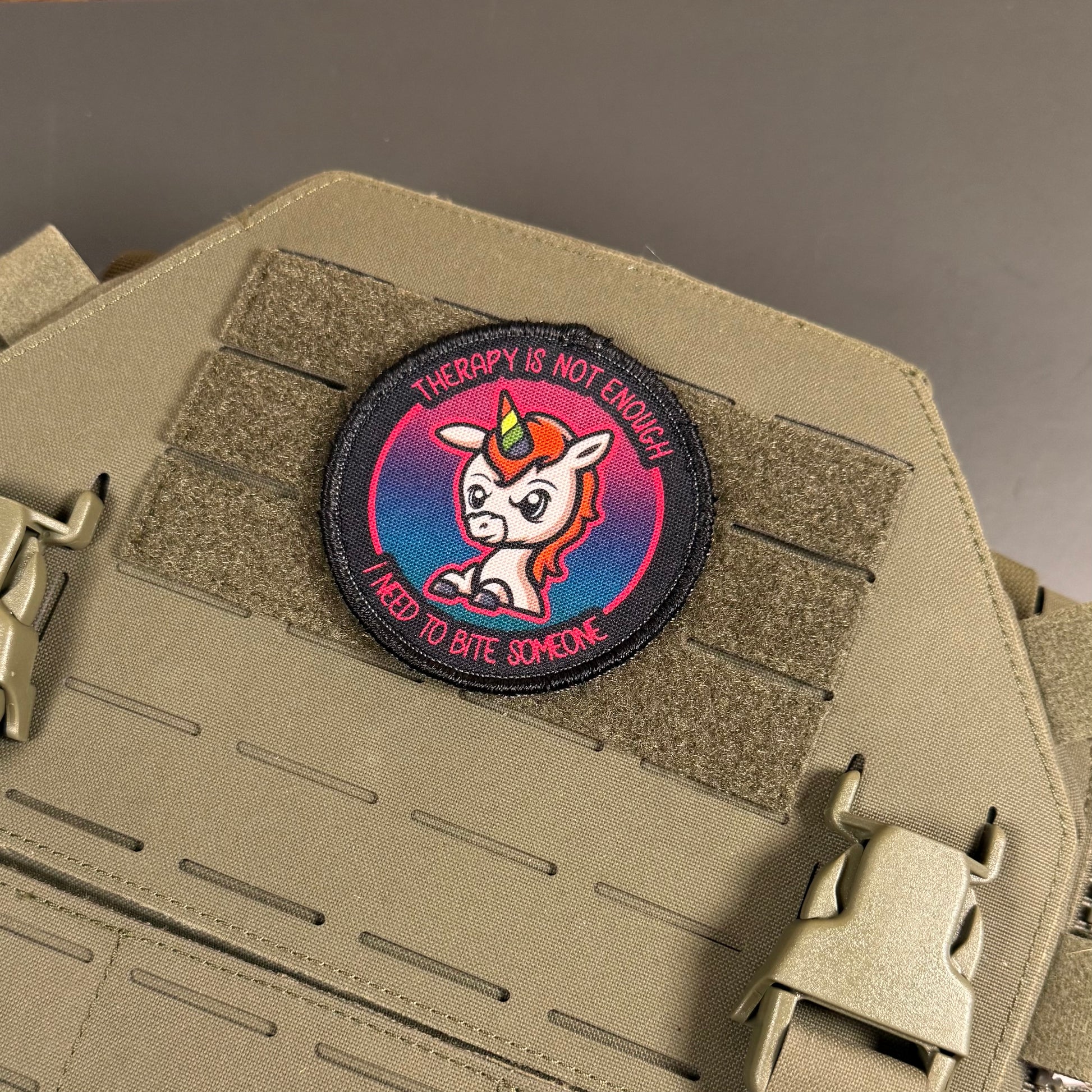 A circular tactical morale patch featuring a cartoon unicorn with the text 'THERAPY IS NOT ENOUGH I NEED TO BITE SOMEONE', displayed on a tan MOLLE tactical vest.