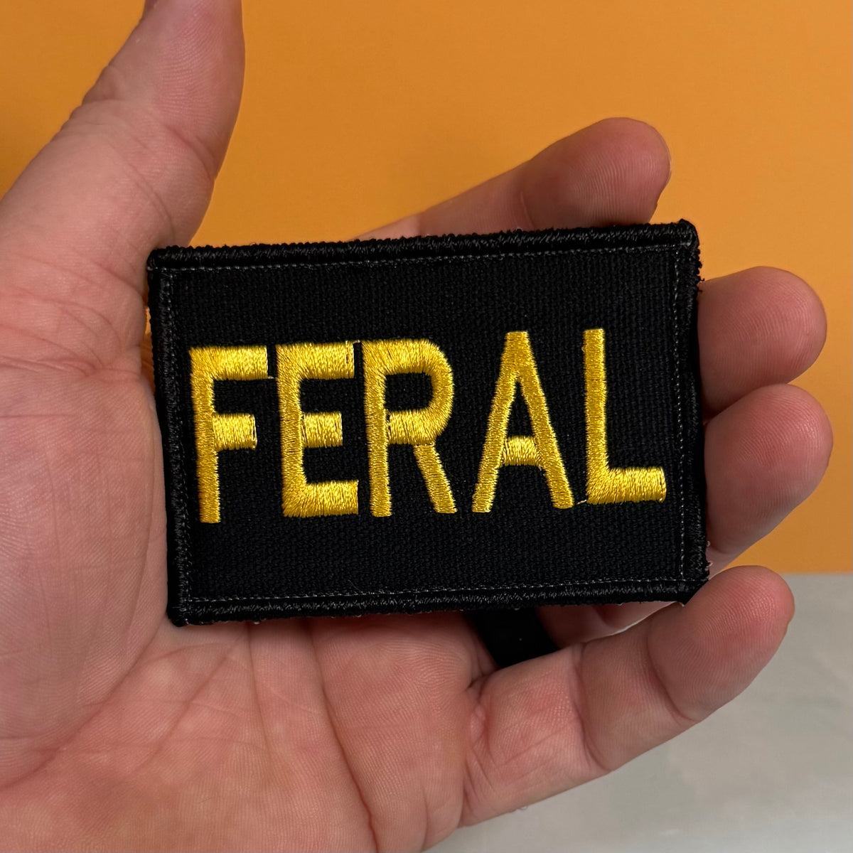 A rectangular black embroidered patch with the word 'FERAL' in bold yellow letters, held in a person's hand against an orange background.