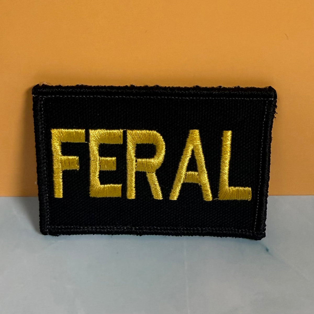 A rectangular black embroidered patch with the word 'FERAL' in bold gold lettering, photographed against a two-tone orange and gray background.