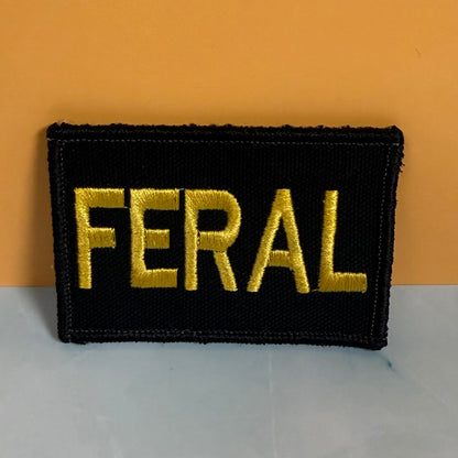 A rectangular black embroidered patch with the word 'FERAL' in bold gold lettering, photographed against a two-tone orange and gray background.