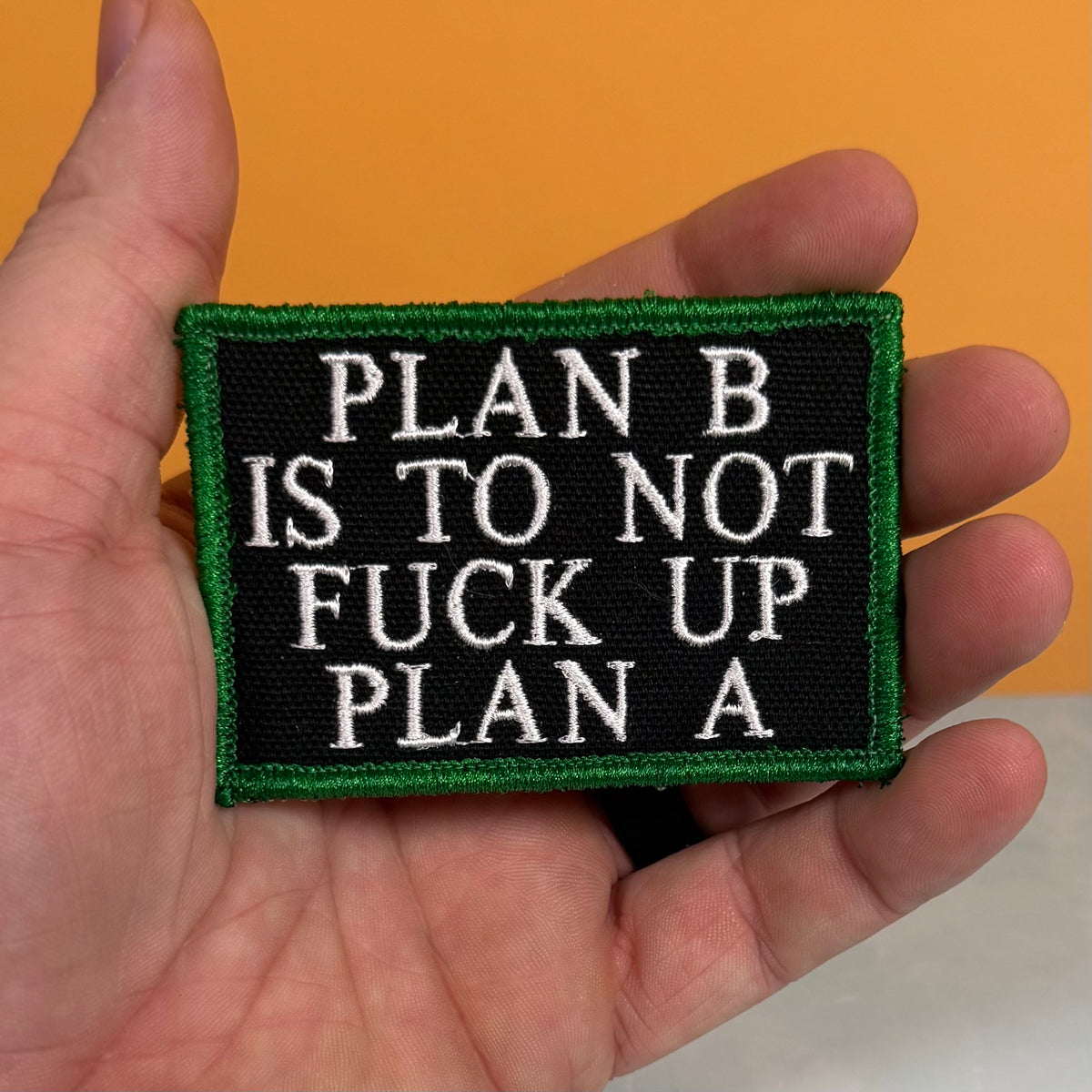 A rectangular embroidered patch held in a hand, featuring a black background with white text reading 'PLAN B IS TO NOT FUCK UP PLAN A' and a green border, against an orange background.