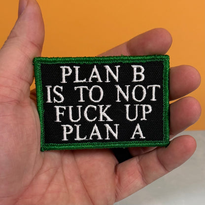 A rectangular embroidered patch held in a hand, featuring a black background with white text reading 'PLAN B IS TO NOT FUCK UP PLAN A' and a green border, against an orange background.