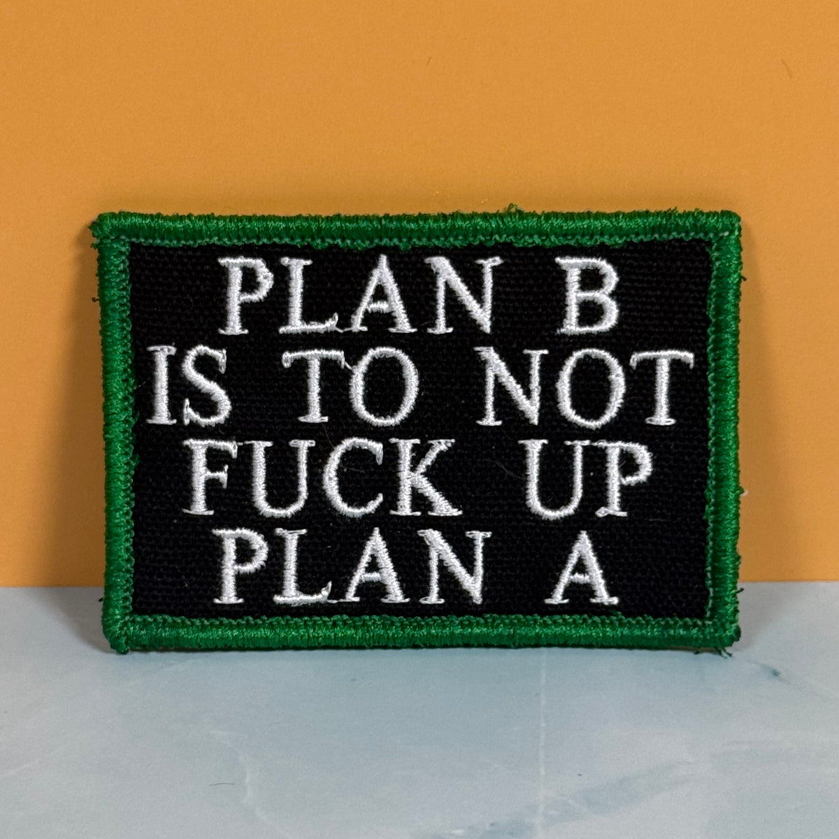 A rectangular embroidered patch with a black center, white text reading 'PLAN B IS TO NOT FUCK UP PLAN A', and a green border, shown against an orange background and light gray surface.