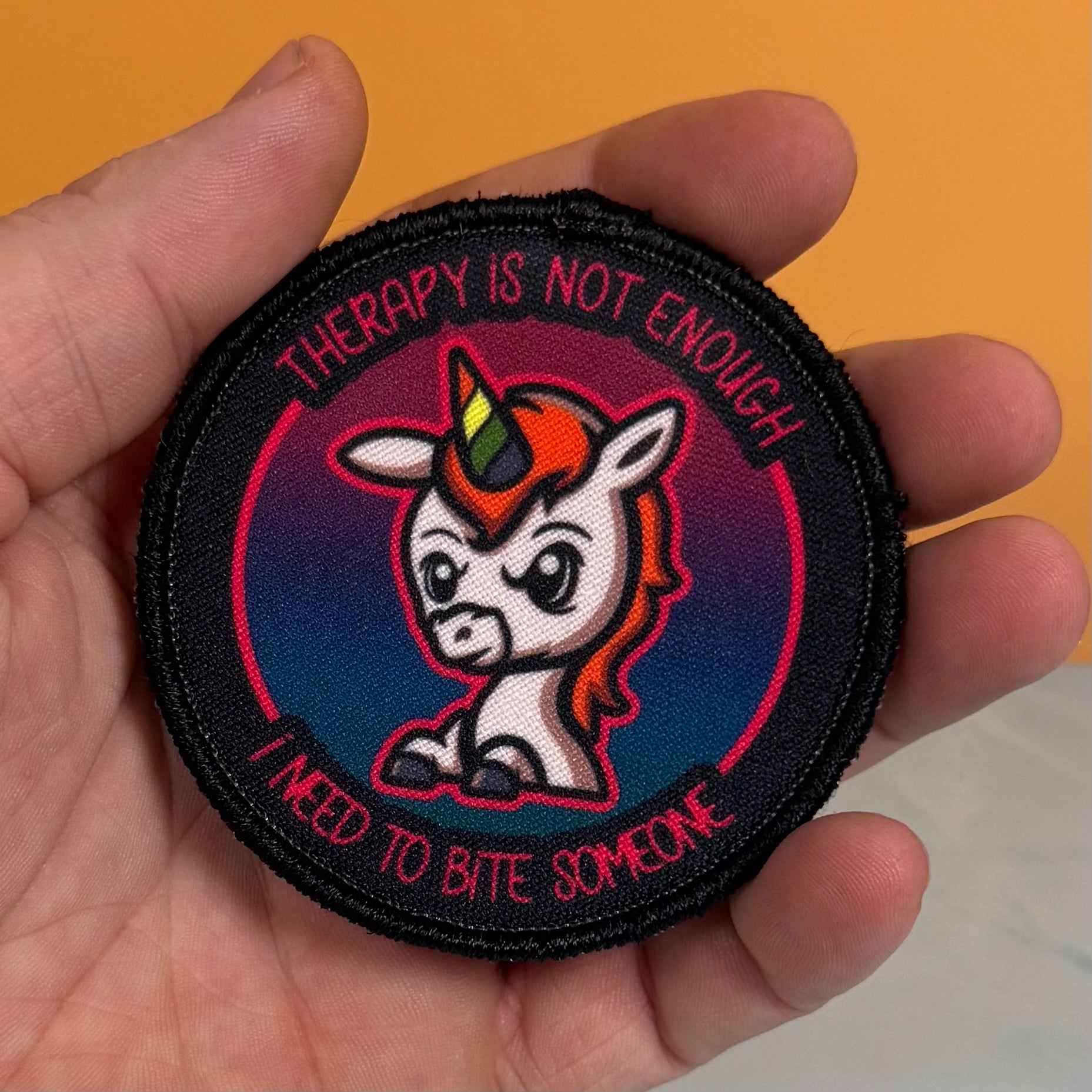 A hand holding a round embroidered patch featuring a cartoon unicorn with a rainbow horn and the text 'THERAPY IS NOT ENOUGH I NEED TO BITE SOMEONE' on a black background.