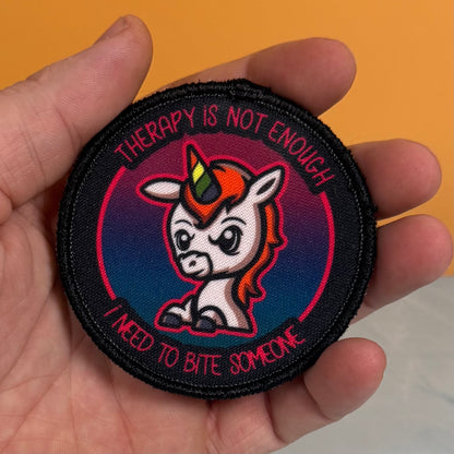 A hand holding a round embroidered patch featuring a cartoon unicorn with a rainbow horn and the text 'THERAPY IS NOT ENOUGH I NEED TO BITE SOMEONE' on a black background.