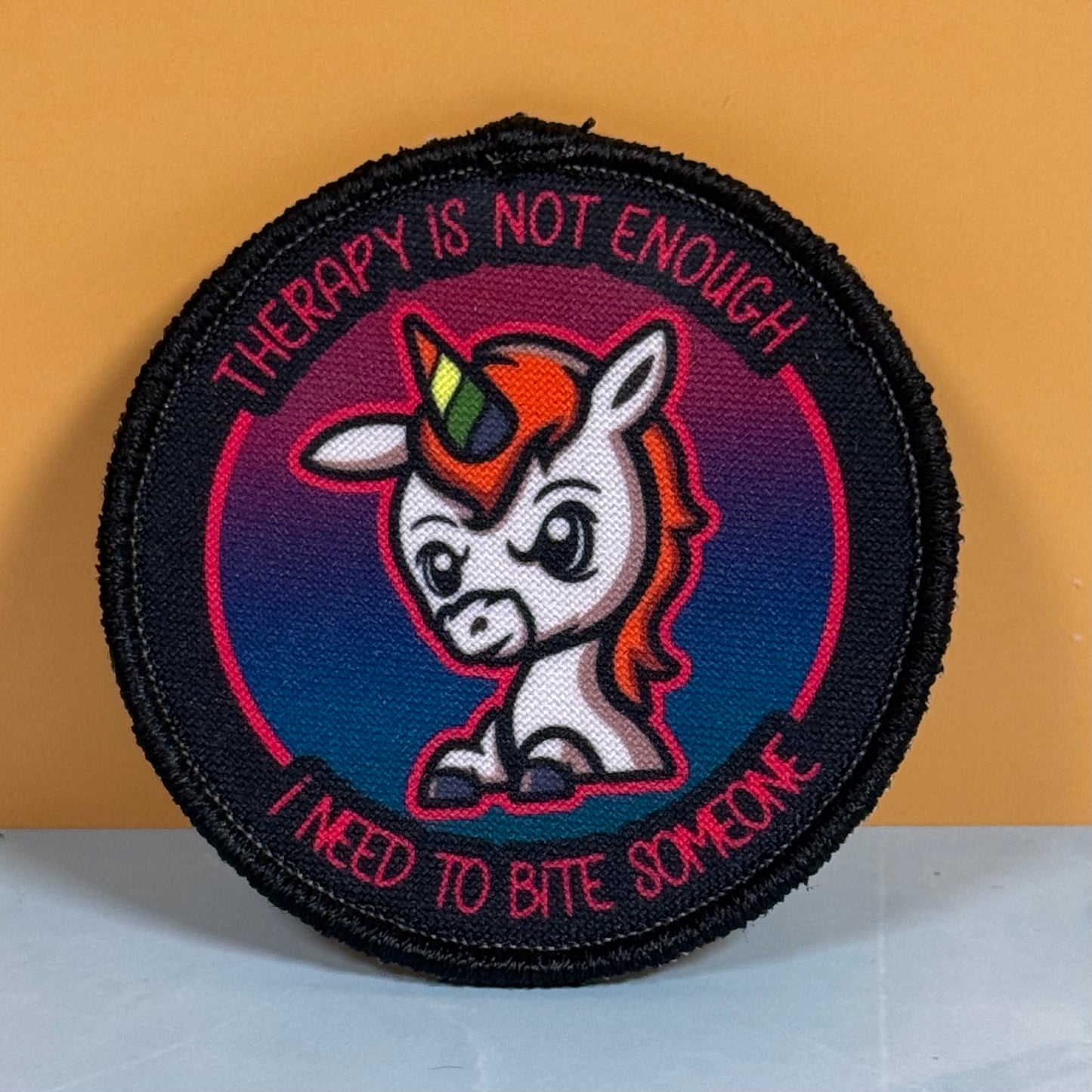 A circular embroidered patch featuring a cartoon unicorn with a rainbow horn, surrounded by the text 'THERAPY IS NOT ENOUGH' and 'I NEED TO BITE SOMEONE', set against a dark blue and pink gradient background with a black stitched border.