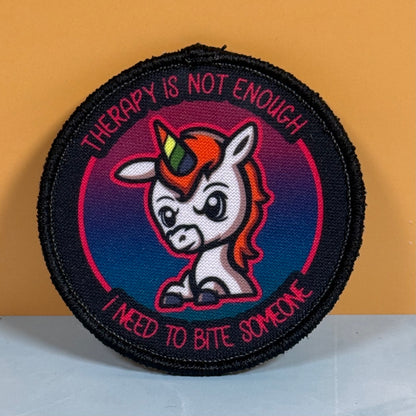 A circular embroidered patch featuring a cartoon unicorn with a rainbow horn, surrounded by the text 'THERAPY IS NOT ENOUGH' and 'I NEED TO BITE SOMEONE', set against a dark blue and pink gradient background with a black stitched border.