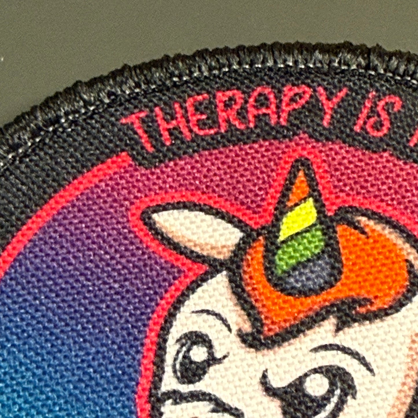 Close-up of an embroidered patch featuring a cartoon unicorn face with a rainbow horn and the text 'THERAPY IS' on a dark background.