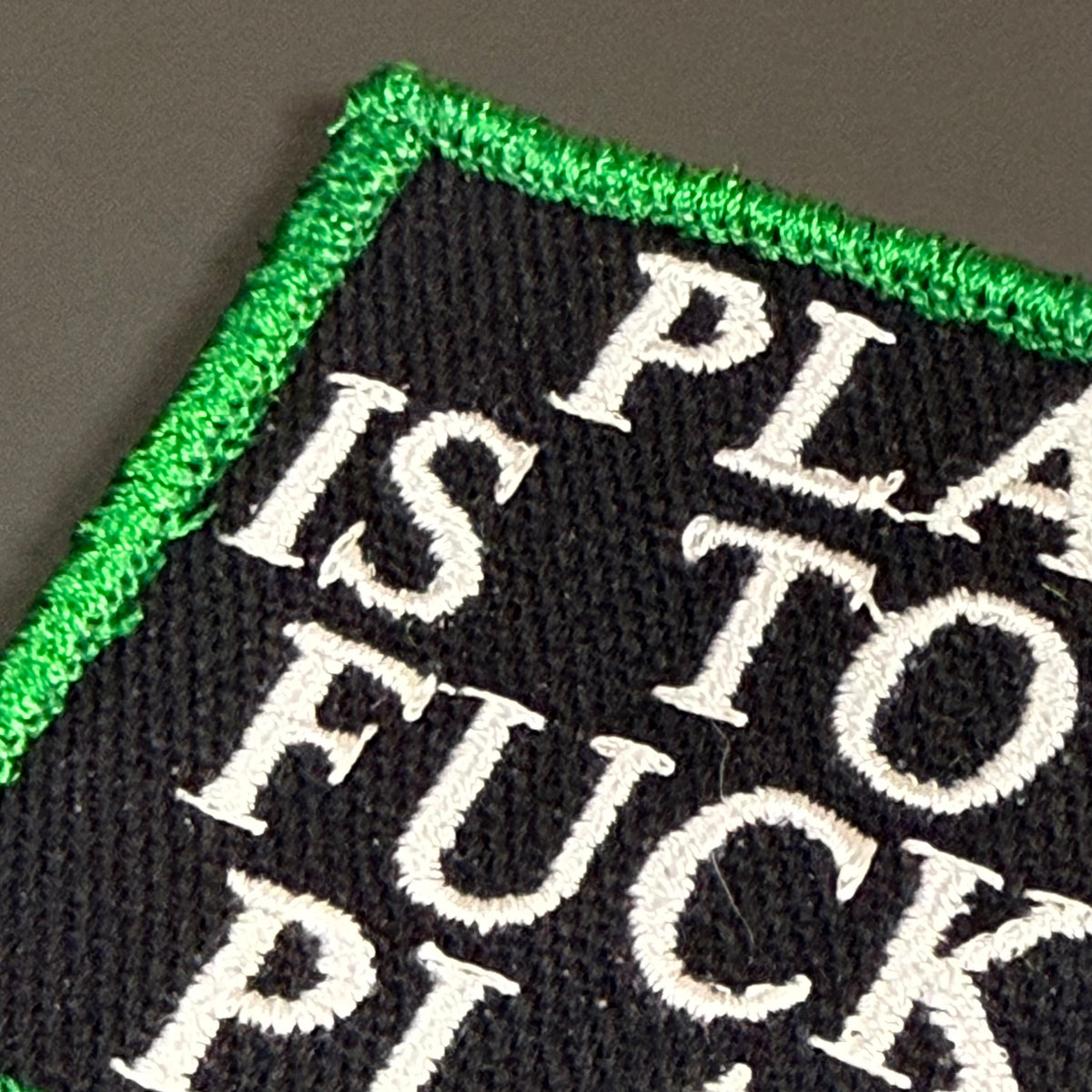 Close-up of an embroidered patch with a black background, white text reading 'PLA IS TO FUCK', and a bright green stitched border on a dark gray surface.
