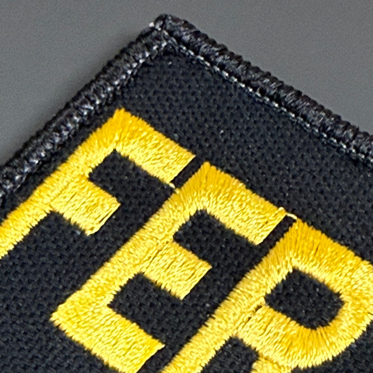 Close-up of a black embroidered patch with bold yellow lettering on a dark gray background.