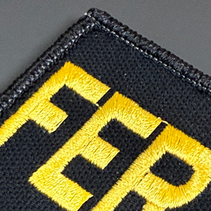 Close-up of a black embroidered patch with bold yellow lettering on a dark gray background.
