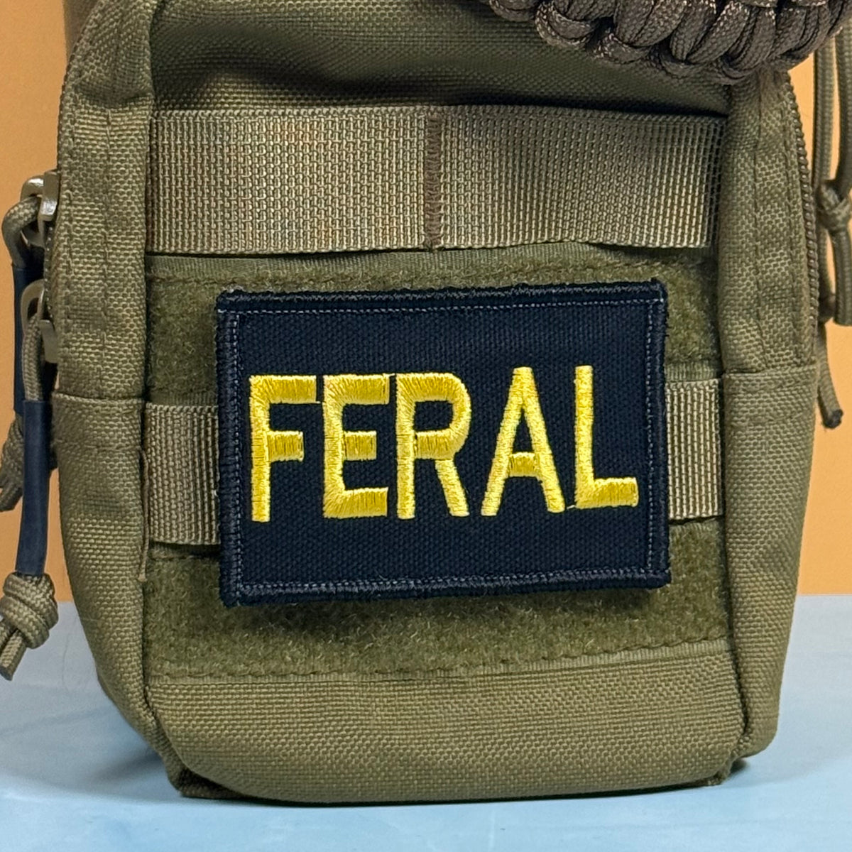 An olive drab tactical pouch featuring a rectangular navy blue patch with the word 'FERAL' embroidered in bright yellow.