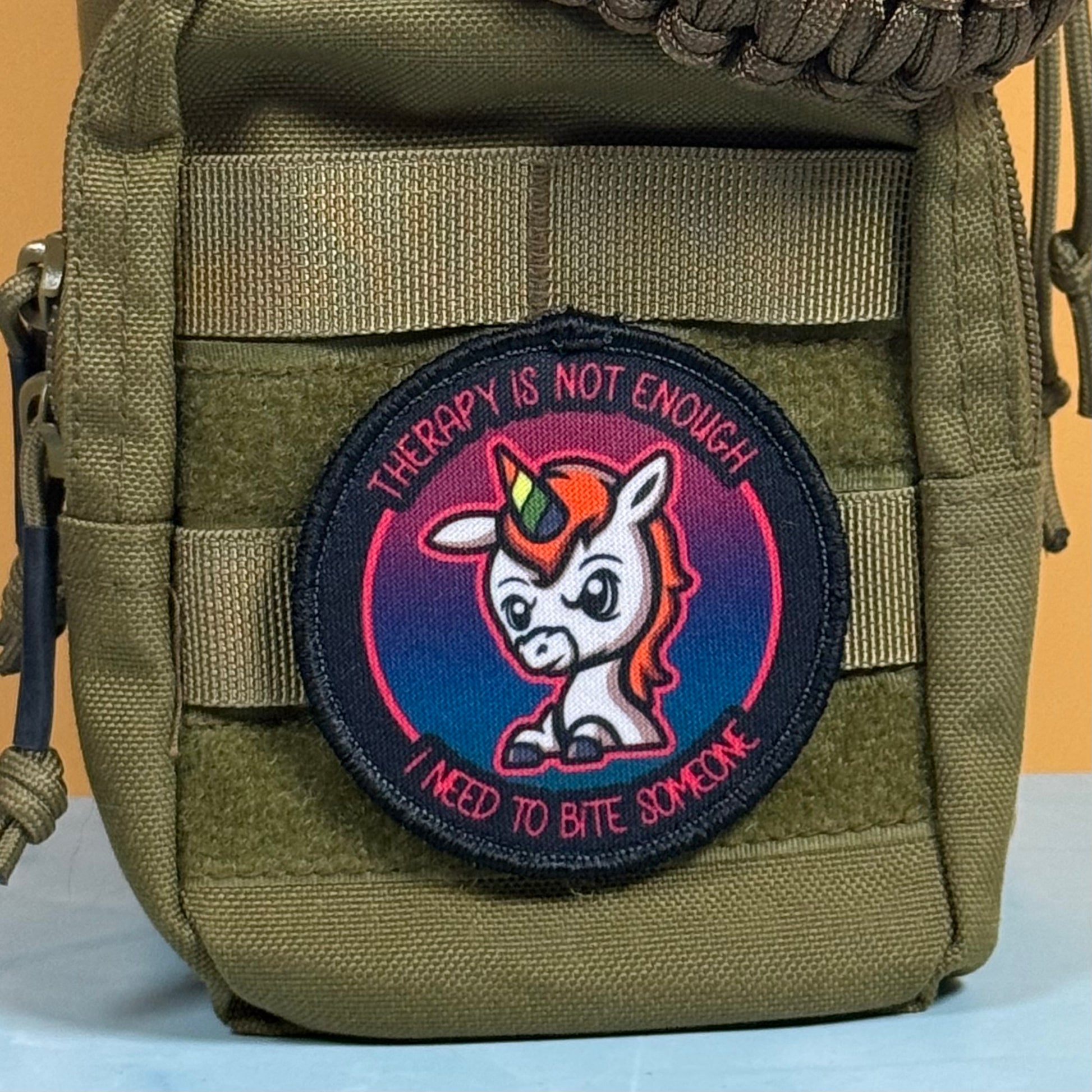 An embroidered circular patch featuring a cartoon unicorn with a rainbow horn and the text 'THERAPY IS NOT ENOUGH I NEED TO BITE SOMEONE', attached to an olive green tactical pouch.