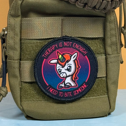 An embroidered circular patch featuring a cartoon unicorn with a rainbow horn and the text 'THERAPY IS NOT ENOUGH I NEED TO BITE SOMEONE', attached to an olive green tactical pouch.