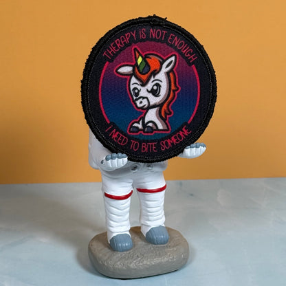 A circular embroidered patch featuring a cartoon unicorn with a rainbow horn and the text 'THERAPY IS NOT ENOUGH I NEED TO BITE SOMEONE', displayed on a small astronaut figurine against a solid orange background.