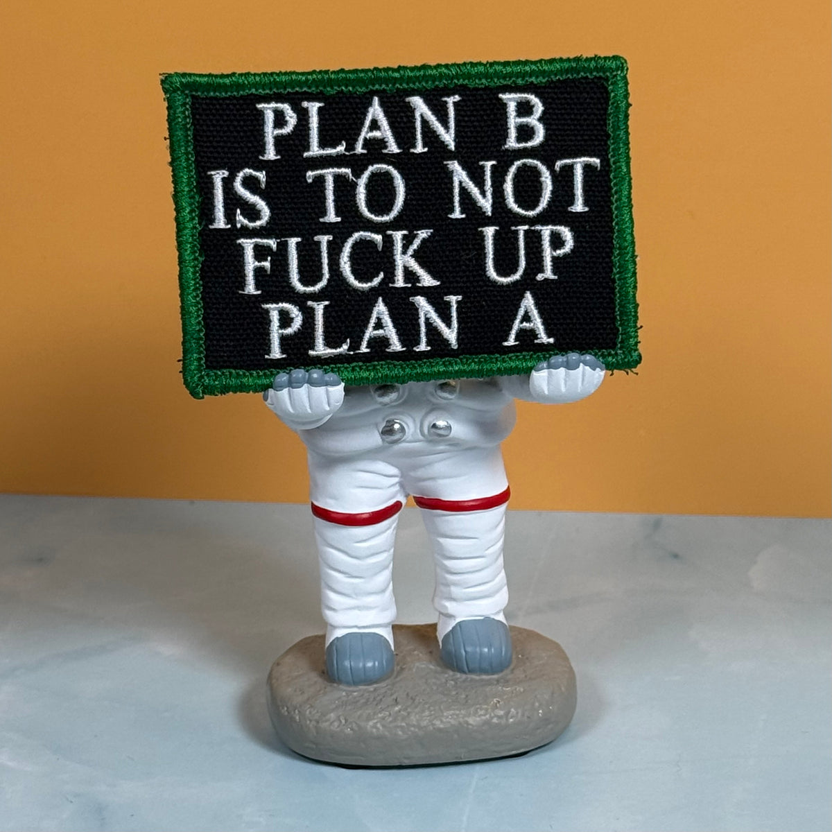 An astronaut figurine holding a black embroidered sign with white text that reads 'PLAN B IS TO NOT FUCK UP PLAN A' and a green border, set against an orange background.
