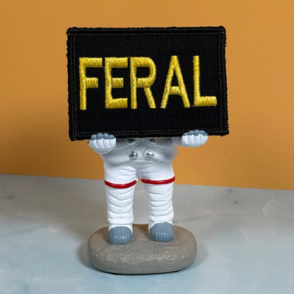 A small astronaut figurine holding a black rectangular patch with the word 'FERAL' in large gold letters. The astronaut is white with red bands and gray boots, standing on a gray base against an orange wall and light gray surface.