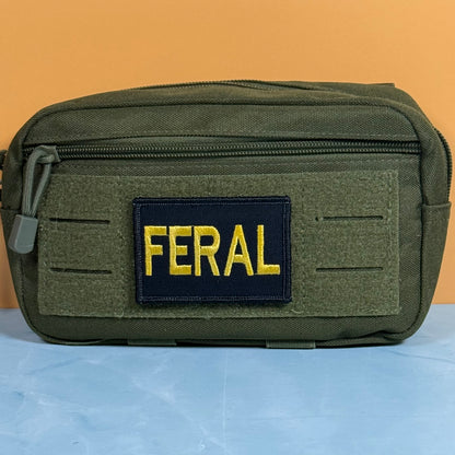 An olive green tactical pouch with a front zippered pocket and a black rectangular patch featuring the word 'FERAL' in yellow embroidery, set against a solid orange background.