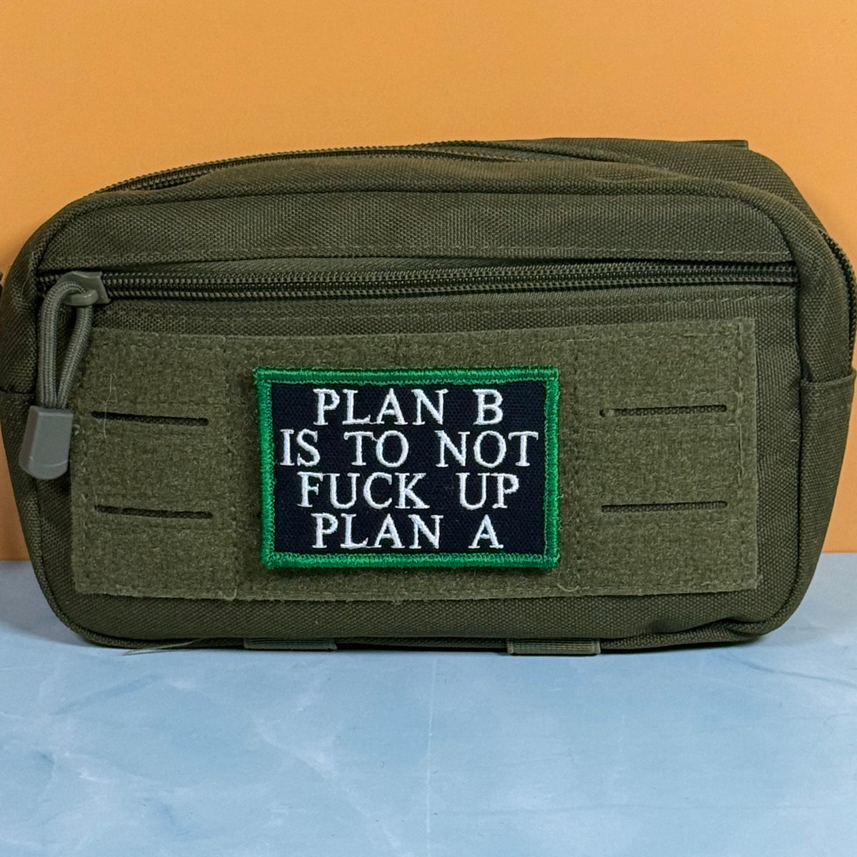 An olive green tactical pouch with a front zipper and a rectangular embroidered patch that reads 'PLAN B IS TO NOT FUCK UP PLAN A' in white text on a black background with a green border, set against an orange and blue background.
