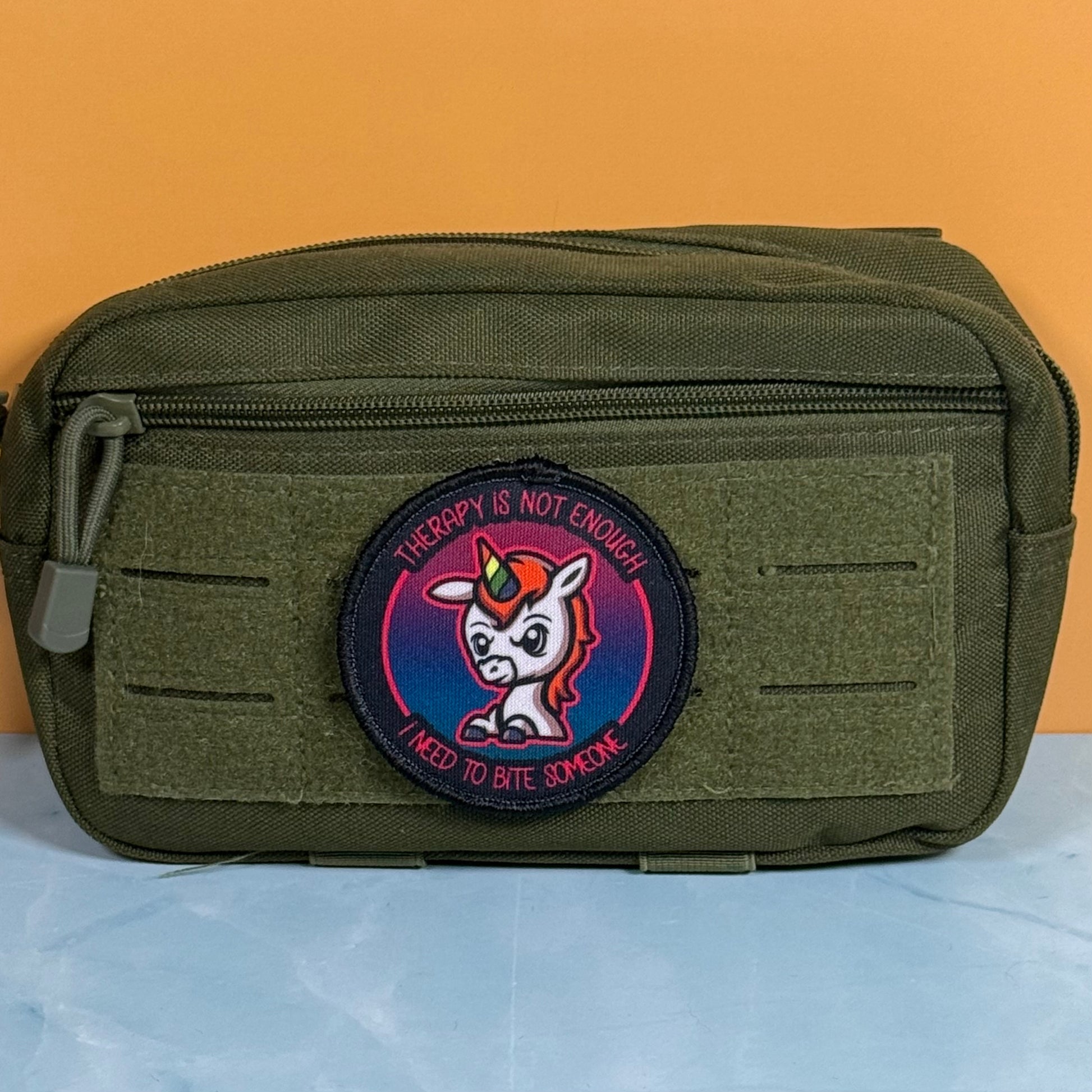 An olive green tactical pouch with a circular embroidered patch featuring a cartoon unicorn and the text 'THERAPY IS NOT ENOUGH I NEED TO BITE SOMEONE', set against a solid orange background.