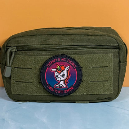 An olive green tactical pouch with a circular embroidered patch featuring a cartoon unicorn and the text 'THERAPY IS NOT ENOUGH I NEED TO BITE SOMEONE', set against a solid orange background.